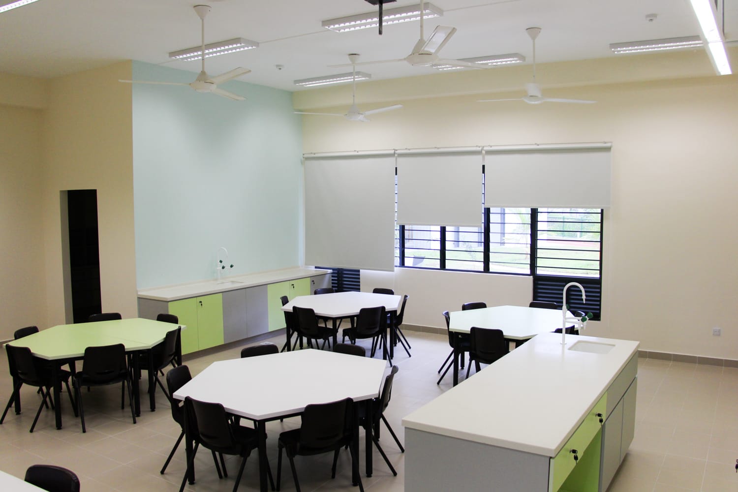 Bright classroom with hexagonal white and green tables, black chairs, ceiling fans, roller blinds, and green cabinets with sinks.